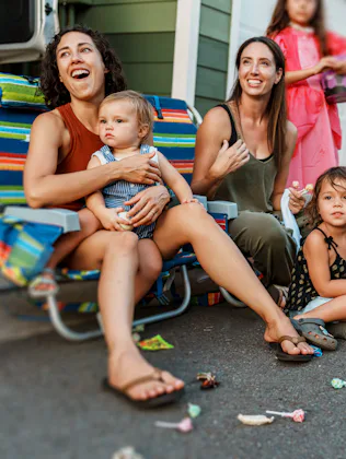 Two women and three young children seated on a curb during a neighborhood event, with costumed child...
