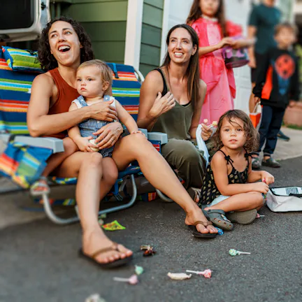 Two women and three young children seated on a curb during a neighborhood event, with costumed child...