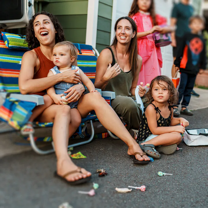Two women and three young children seated on a curb during a neighborhood event, with costumed child...