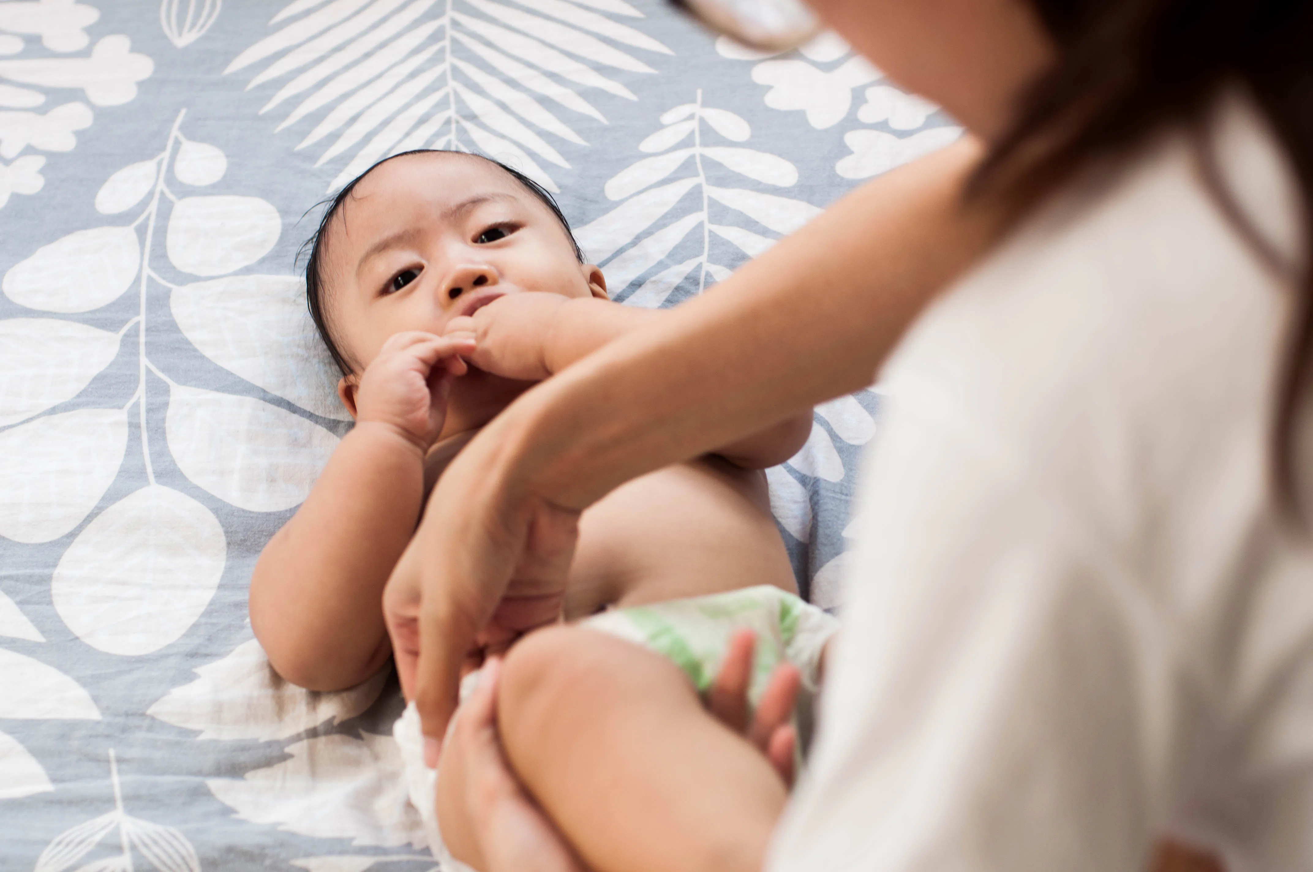 A caregiver adjusts a diaper on a reclining infant on a patterned bedspread, the baby looking up whi...