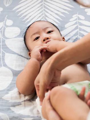 A caregiver adjusts a diaper on a reclining infant on a patterned bedspread, the baby looking up whi...