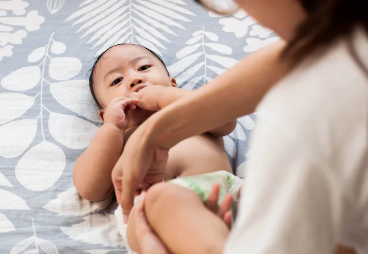 A caregiver adjusts a diaper on a reclining infant on a patterned bedspread, the baby looking up whi...