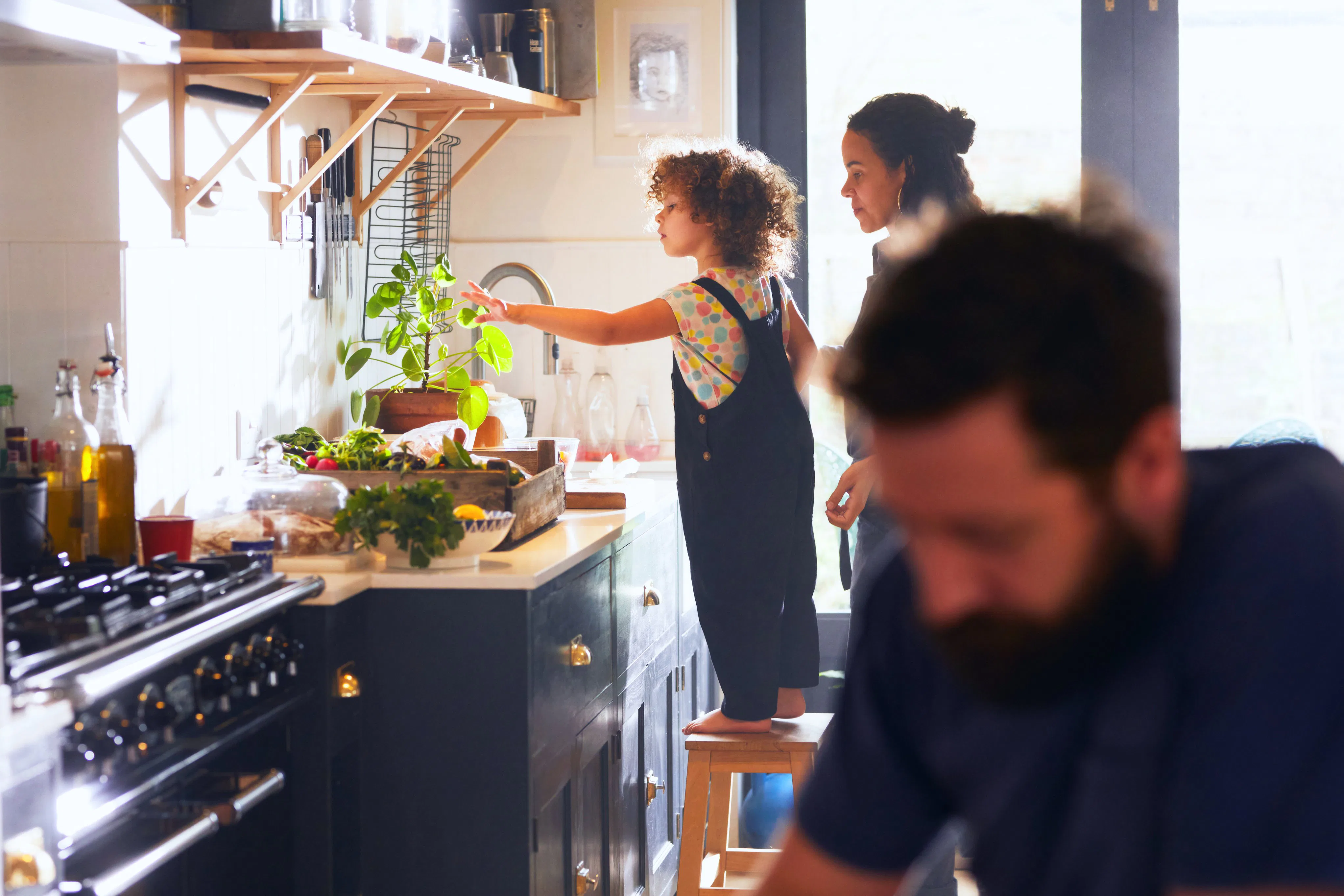 Child standing on a stool at a kitchen counter reaching toward a plant while an adult watches; a bea...