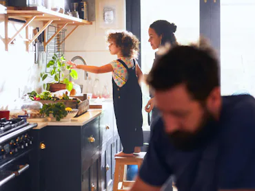 Child standing on a stool at a kitchen counter reaching toward a plant while an adult watches; a bea...