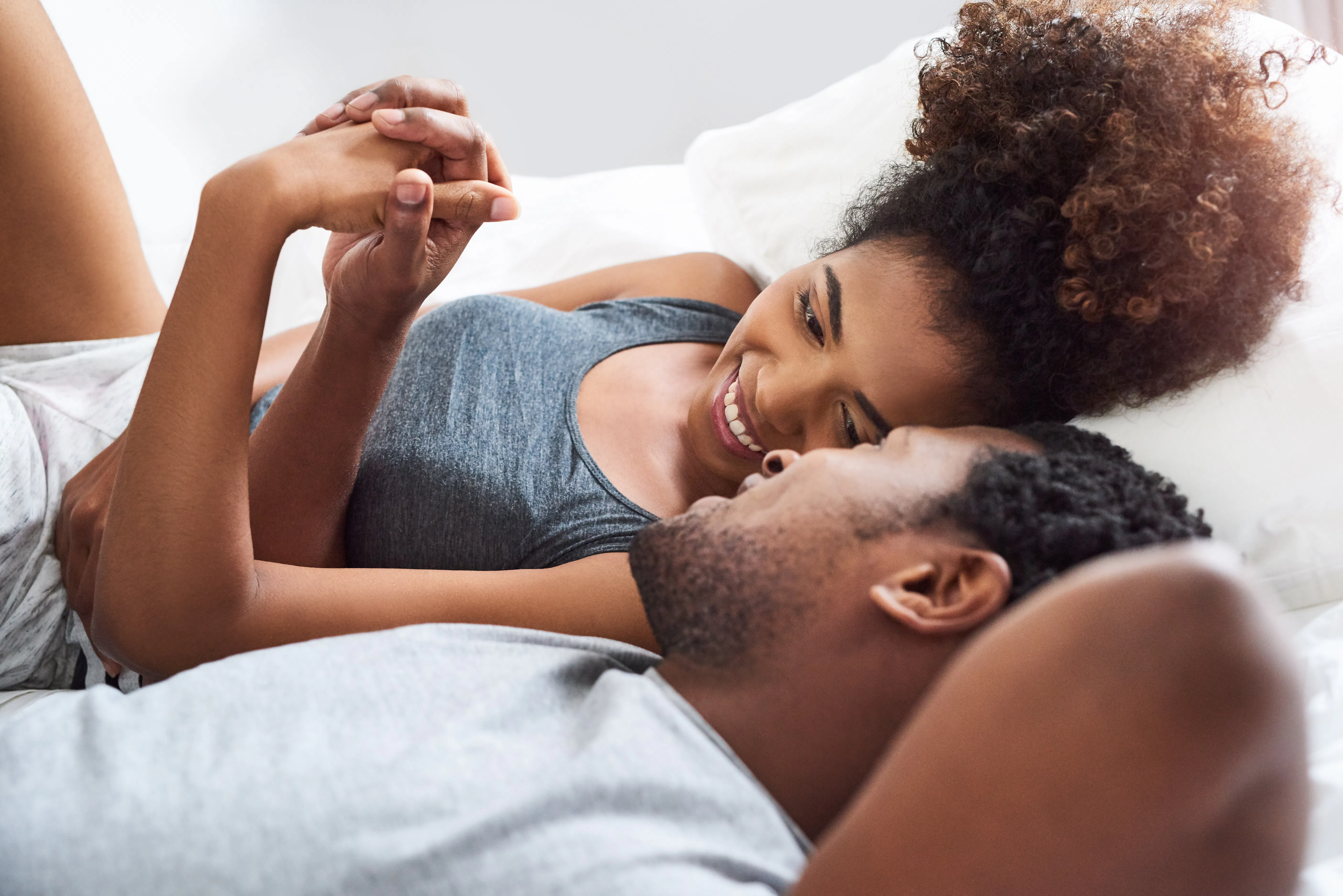 Young Black couple lying on a bed facing each other, smiling and holding hands, woman propped on elb...