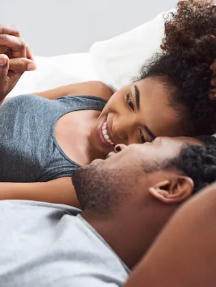Young Black couple lying on a bed facing each other, smiling and holding hands, woman propped on elb...