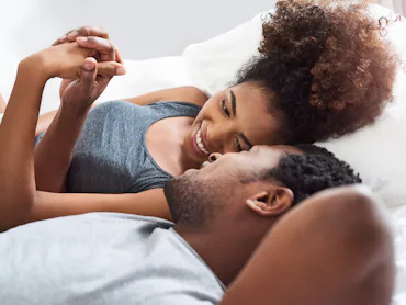 Young Black couple lying on a bed facing each other, smiling and holding hands, woman propped on elb...