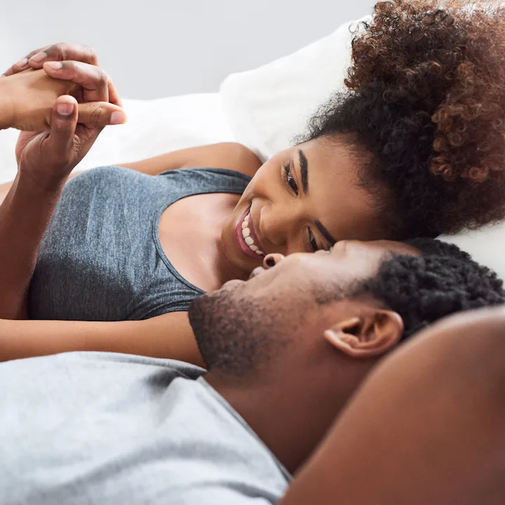 Young Black couple lying on a bed facing each other, smiling and holding hands, woman propped on elb...