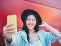 A happy woman in a black hat takes a selfie in front of a red wall. 