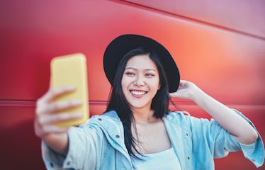 A happy woman in a black hat takes a selfie in front of a red wall.