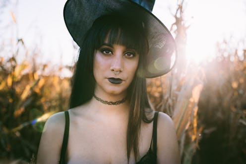 Halloween costume witch girl portrait in a cornfield at sunset. Beautiful serious young woman in wit...