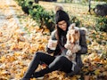 A young woman sits with her dog while drinking a latte in the fall in a pile of yellow leaves.
