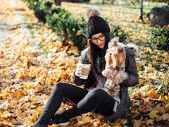 A young woman sits with her dog while drinking a latte in the fall in a pile of yellow leaves.