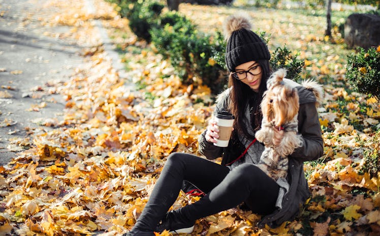 A young woman sits with her dog while drinking a latte in the fall in a pile of yellow leaves.