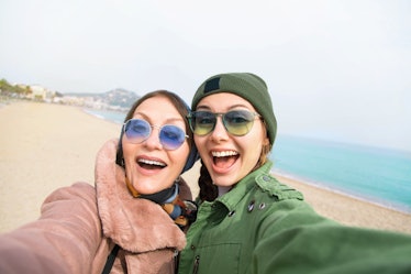 Daughter poses with mom for selfie on the beach while traveling.
