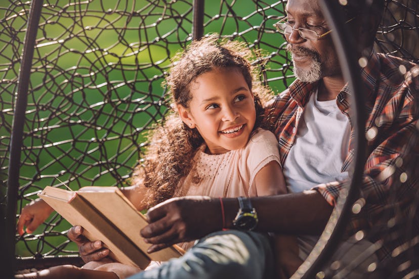 smiling african american granddaughter reading book while sitting in swinging hanging chair with gra...