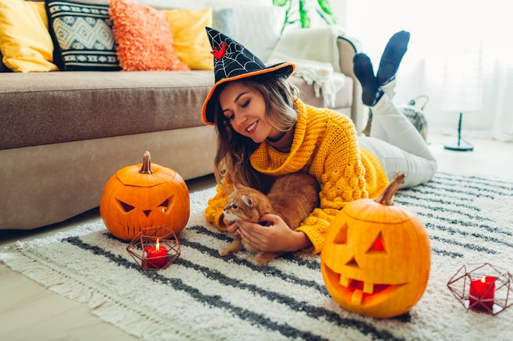 Halloween jack-o-lantern pumpkins. Woman in hat playing with cat lying on carpet at home decorated w...