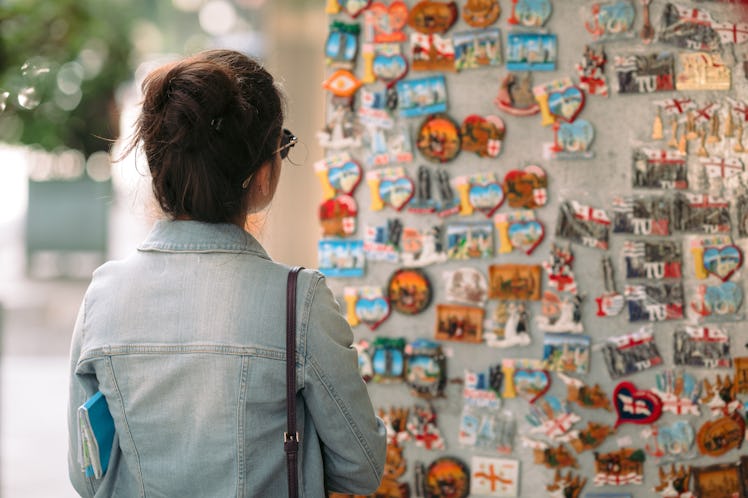 A brunette woman in a denim jacket looks at magnets in a souvenir shop while traveling.