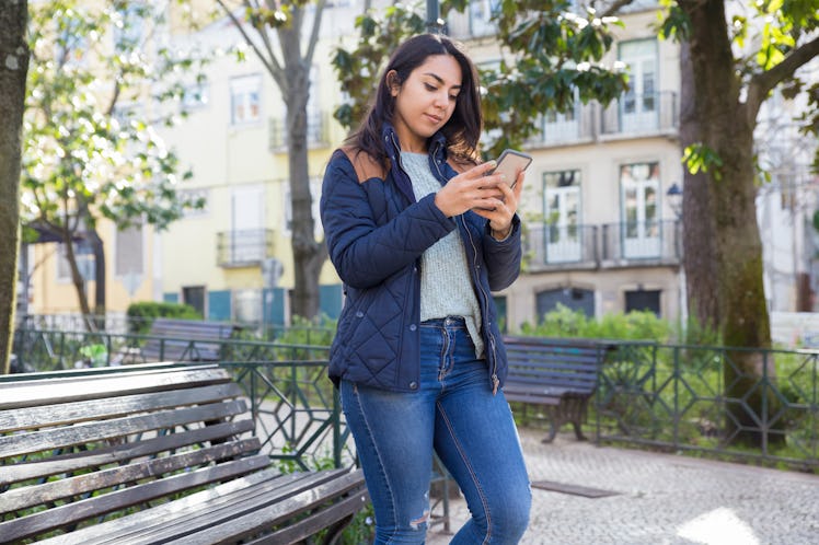 Serious woman using smartphone and standing outdoors. Pretty young lady wearing jacket with trees an...