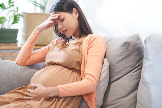 Pregnant woman experiencing a headache holds her hadn to her head while sitting on the couch