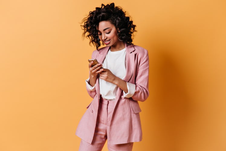 Smiling girl in pink jacket texting message. Glad young woman in suit holding smartphone.