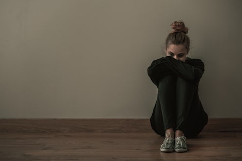 Young woman with anxiety disorder wearing dark clothes sitting on the floor, copy space on empty wal...