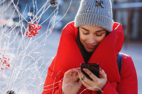close up portrait of happy woman in red winter down jacket and knitted grey hat types text of sms or...