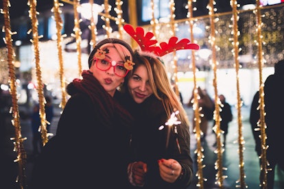 Two women dressed all festive standing in front of a row of Christmas lights outside in the winter.