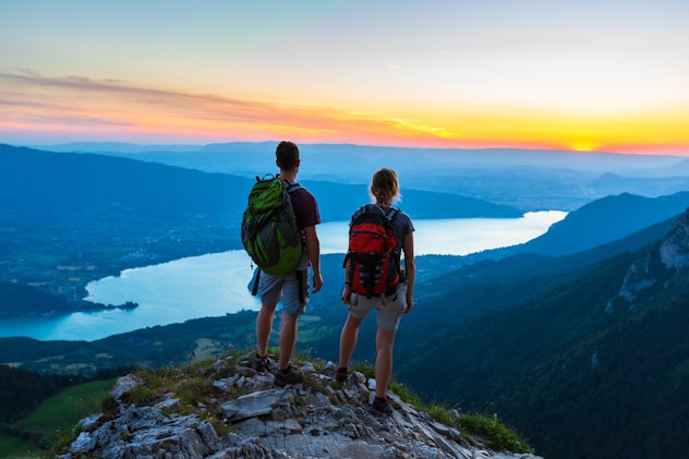 Hikers enjoying scenic view of valley with lake at sunset, couple enjoying summer outdoor trek in mo...