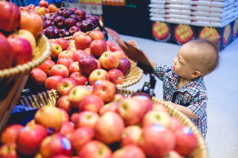 A toddler stands at an apple display at a grocery store, holding fruit and smiling. 