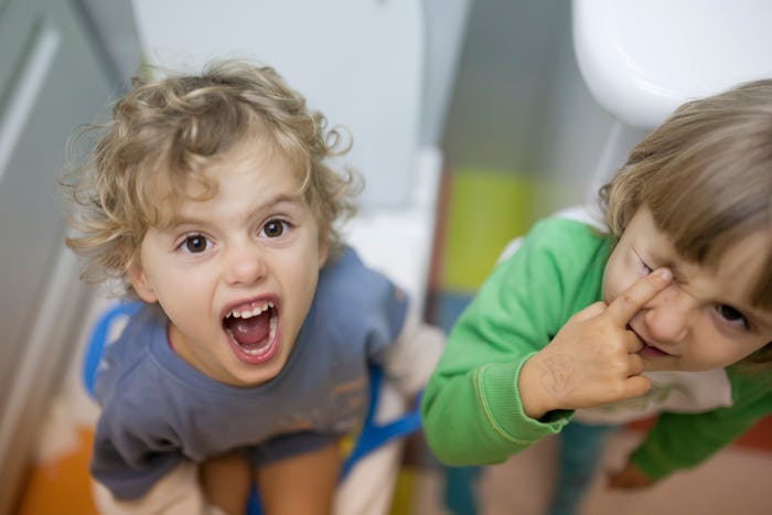 Angry little girl sits on toilet training seat, screaming at camera, while her twin sister stands ne...