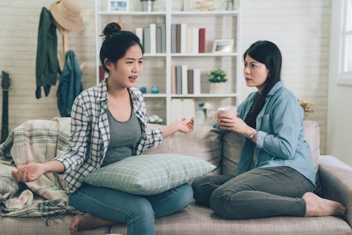 Two female friends sitting on sofa chatting talking and drinking tea or coffee spending time togethe...