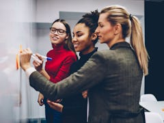 Smiling multiracial women colleagues standing near flip chart for drawing graphic of productivity co...