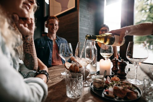 Man hand pouring white wine from the bottle into glasses with friends sitting around the table. Grou...