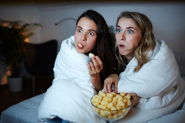 Two girls eating corn rolls from a glass bowl while watching a movie at night in bed.