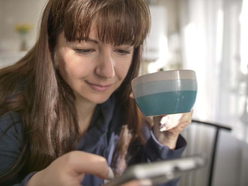 Young woman holding a coffee mug and using the phone in the kitchen.