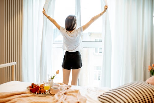Young woman stretching near the window, having a good morning at the cozy bedroom with breakfast at ...