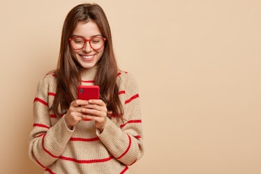 Indoor photo of satisfied teenage girl texts on cellular, reads interesting article online, wears ca...