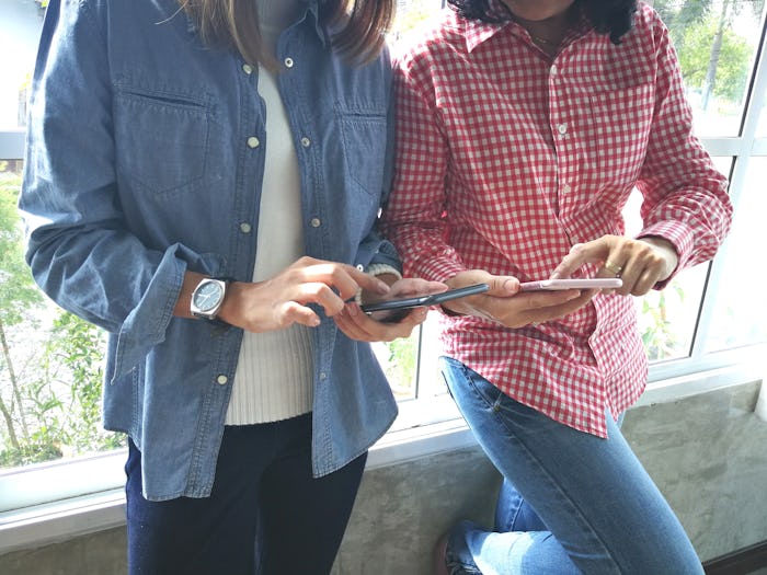 Two women leaning against a window, texting on their phones. 