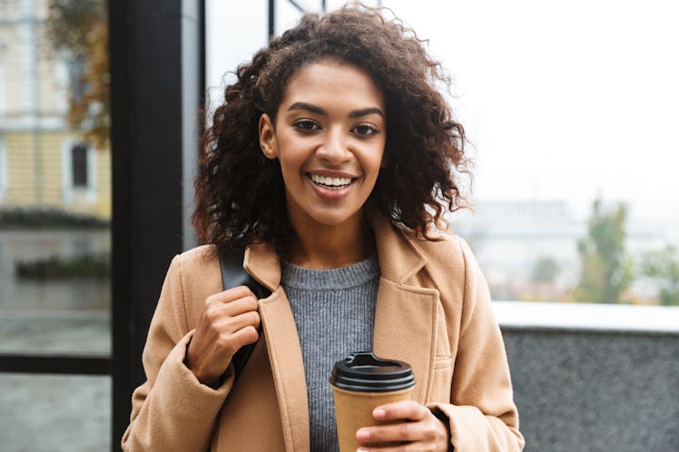 A young woman with a tan coat walks outdoors and holds a to-go coffee cup in her hand, for which she...