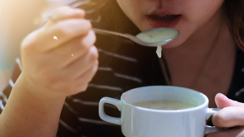 Close up and selective focus of asian woman eating mushroom soup with silver spoon from white cup .