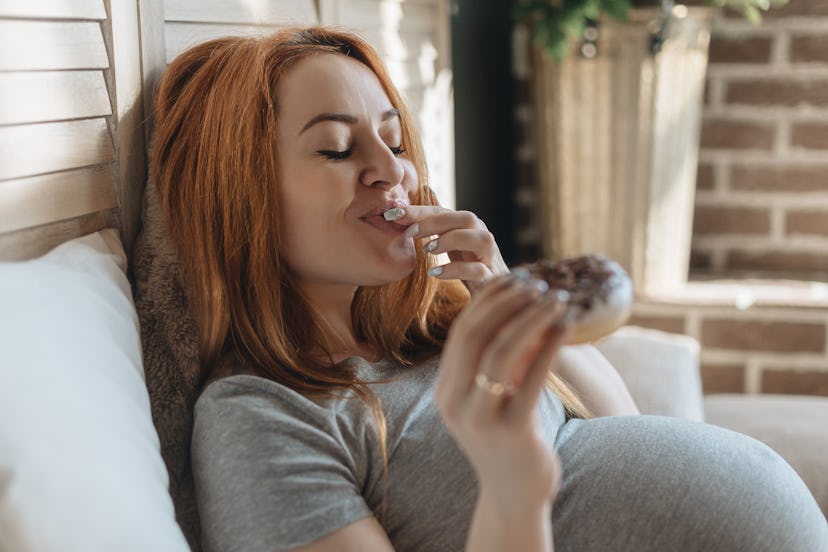Pregnant woman eating tasty donuts while sitting on bed and smiling. Yummy mom. Enjoying food being ...