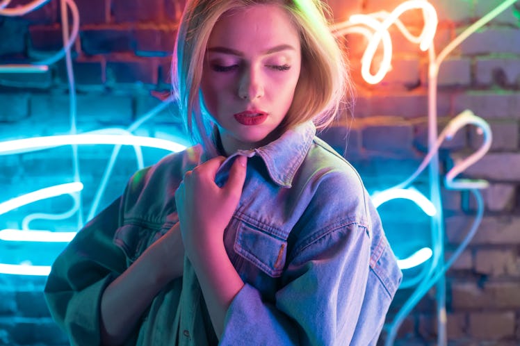 Attractive young woman wearing stylish trendy denim jacket standing near brick wall illuminated with...
