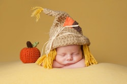 Four week old newborn baby boy wearing a crocheted scarecrow hat. He is sleeping on a gold blanket n...