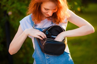 A red-haired girl looks through her black fanny pack that has multiple zippers, which makes it one o...