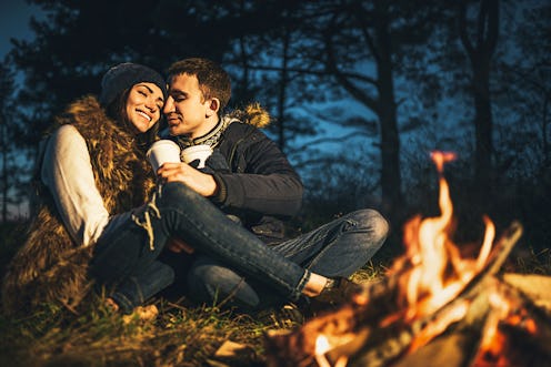 Pretty young couple drinking hot beverage in the forest near bonfire