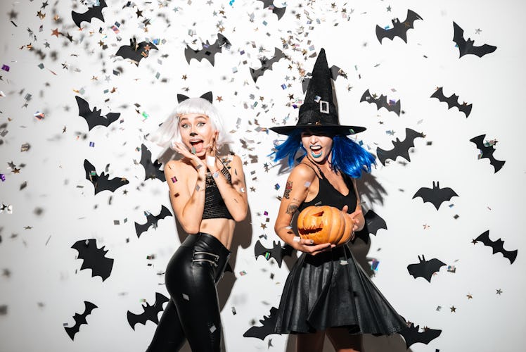 Two women dressed up for Halloween — one as a cat and one as a witch — smile as confetti falls aroun...