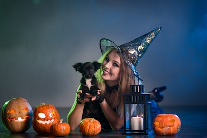 A woman dressed in a witch costume holding her dog, surrounded by pumpkins on Halloween.