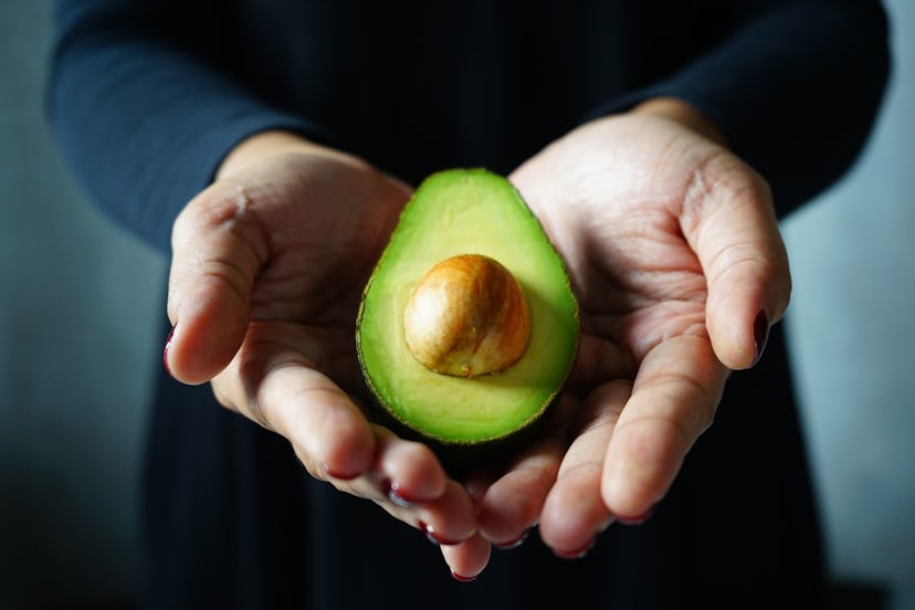 Fresh cut avocado in hands of woman in black dress with blurred dark grey background. Being vegan is...