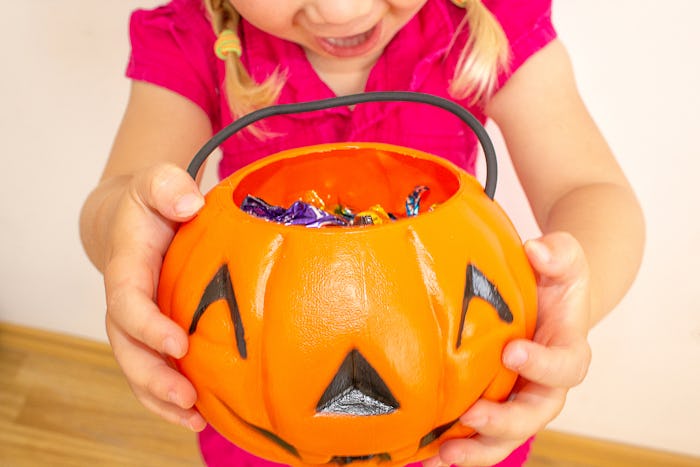 A little girl holds a pumpkin with candy in her hands and stretches it to get even more candies for ...